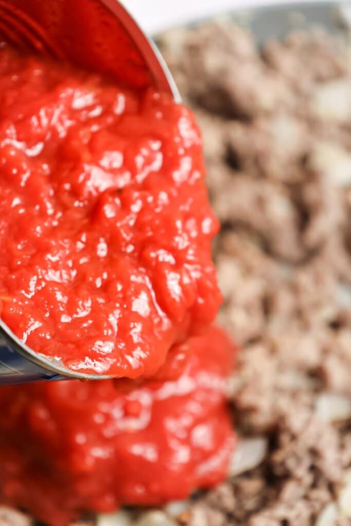 Crushed tomatoes being poured into ground beef.