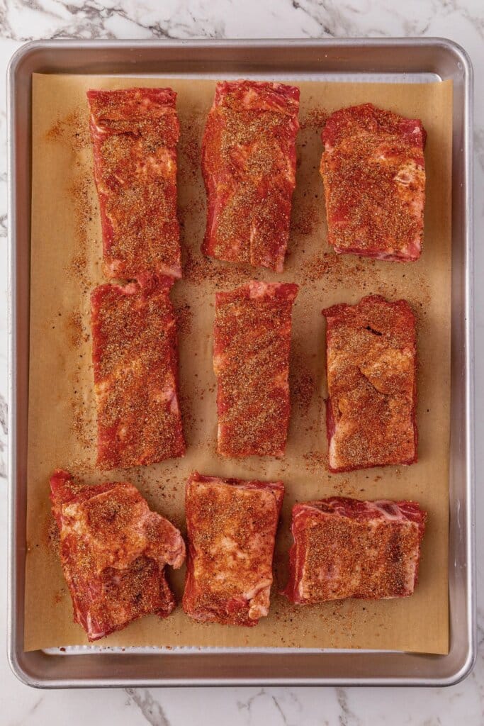 Meat being seasoned on a baking sheet.