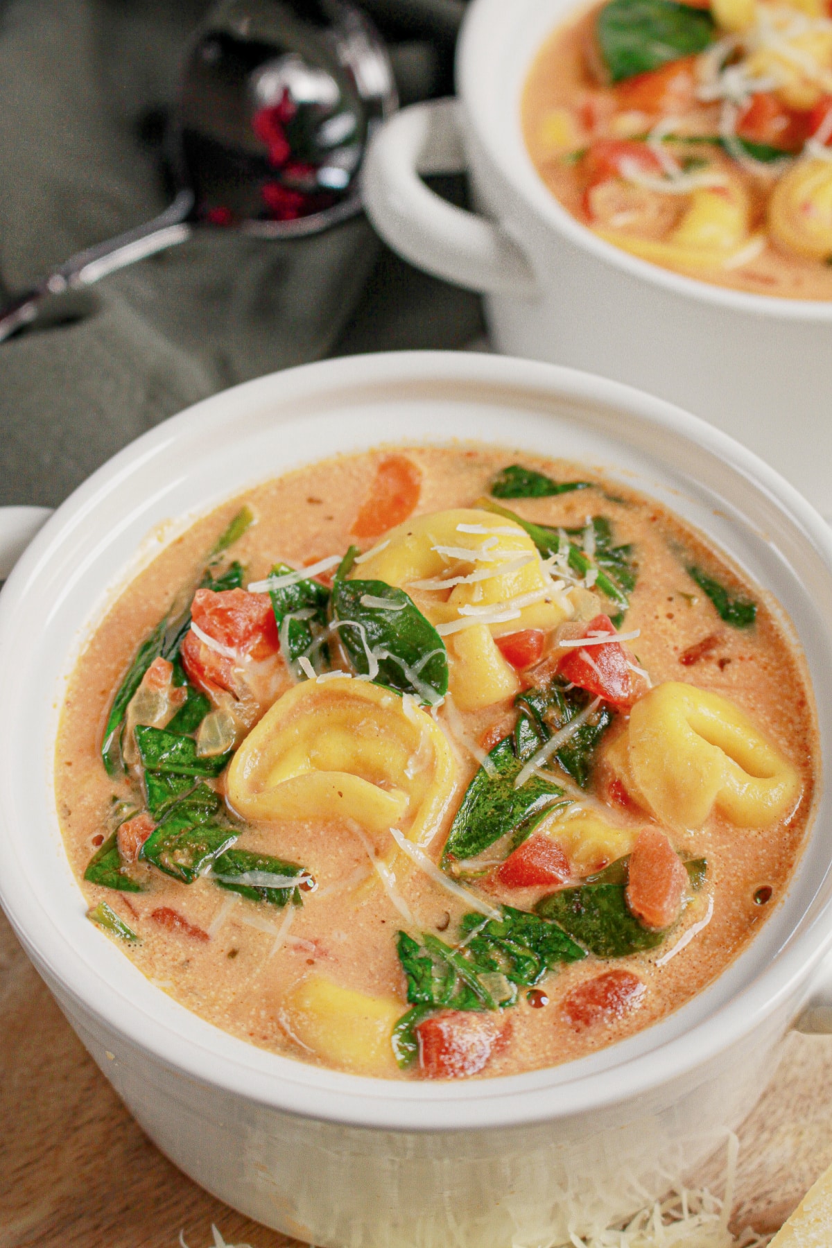 A bowl of creamy tortellini soup with spinach, diced tomatoes, and shredded cheese, served in a white dish.
