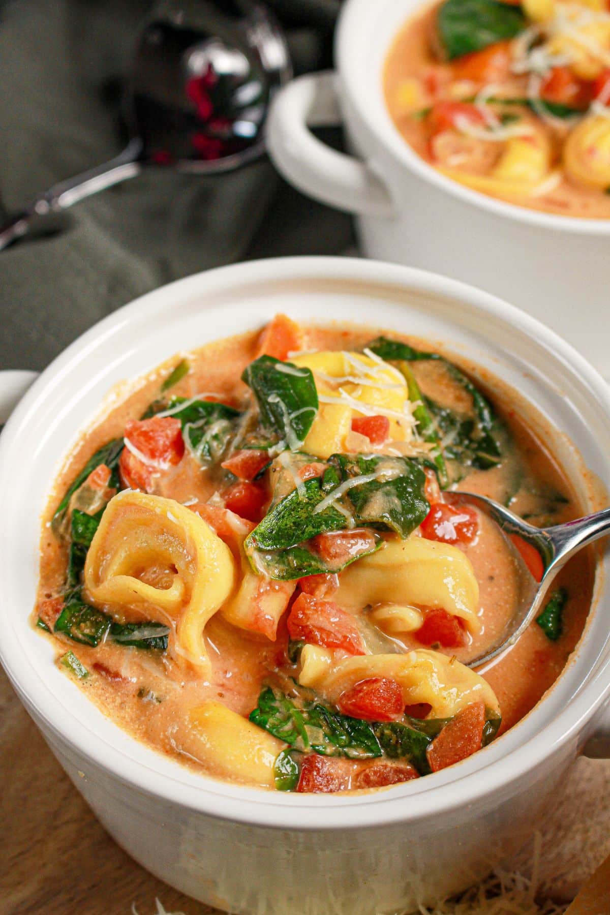 A white bowl of creamy tortellini soup with spinach, tomatoes, and shredded cheese, with a spoon in the bowl.