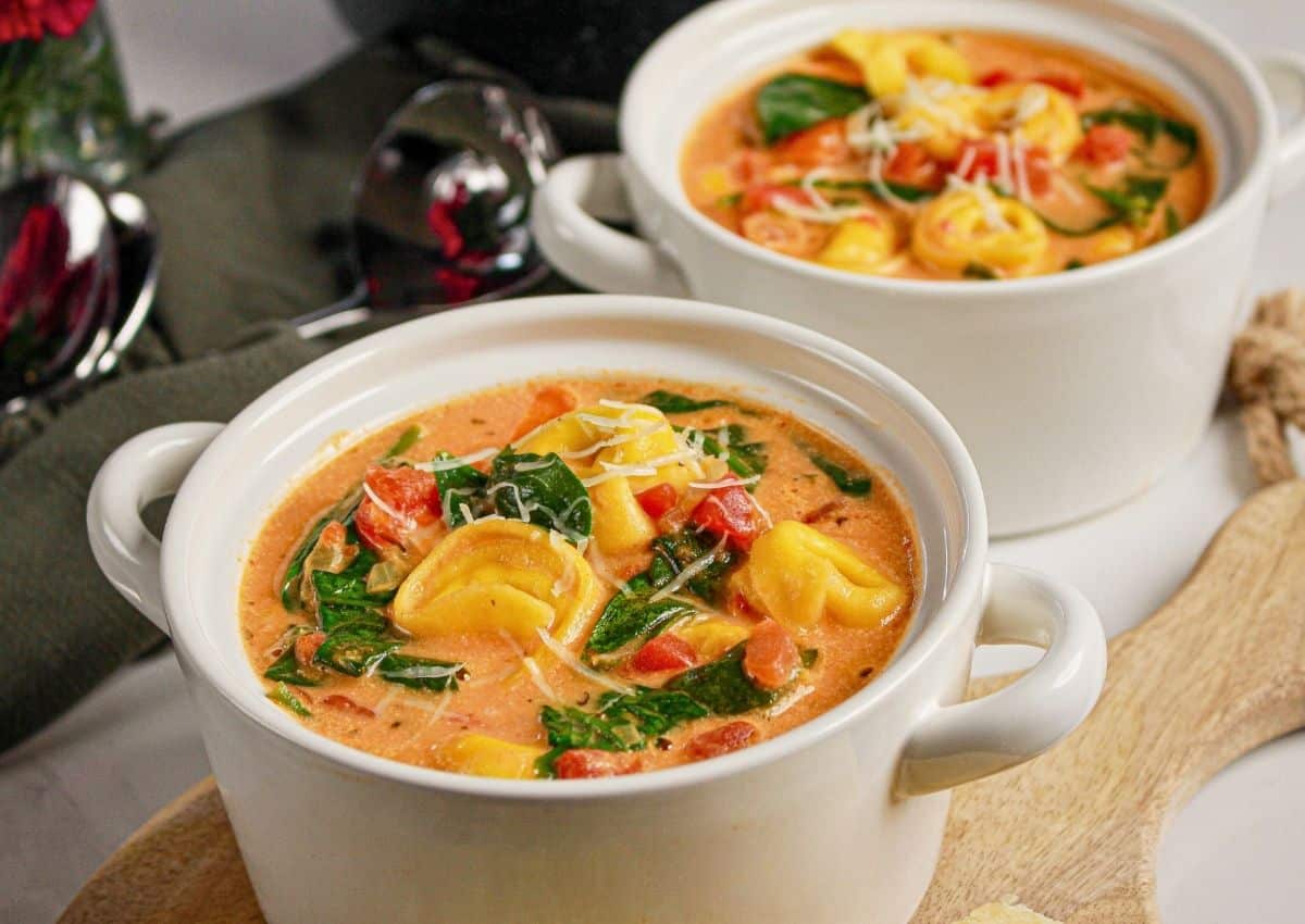 Two white bowls filled with creamy tortellini soup with spinach, tomatoes, and shredded cheese on top, placed on a wooden board.