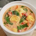 A bowl of creamy tortellini soup with spinach, tomatoes, and grated cheese in a white ceramic dish.