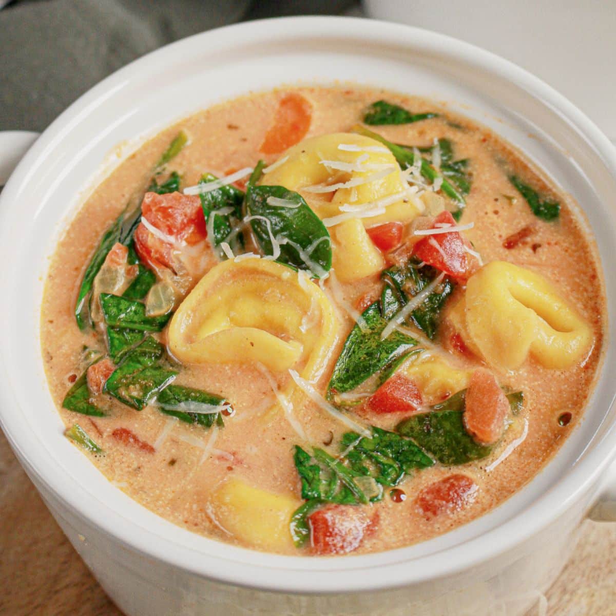 A bowl of creamy tortellini soup with spinach, tomatoes, and grated cheese in a white ceramic dish.