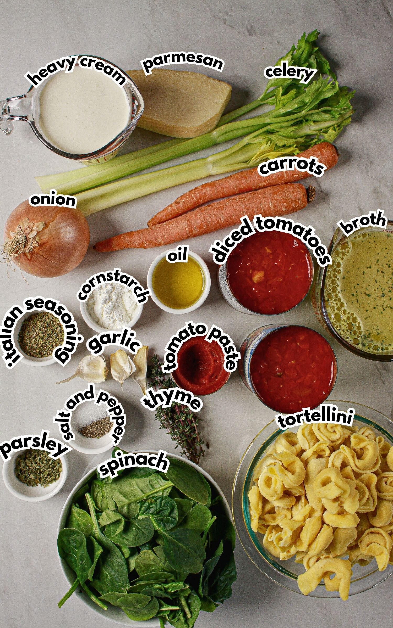 Ingredients for a tortellini soup recipe arranged on a countertop, including tortellini, spinach, carrots, celery, onion, garlic, broth, diced tomatoes, tomato paste, cream, parmesan, and seasonings.