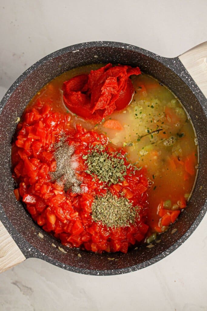 A pot containing chopped tomatoes, tomato paste, broth, herbs, and spices on a light countertop, ready to be cooked.