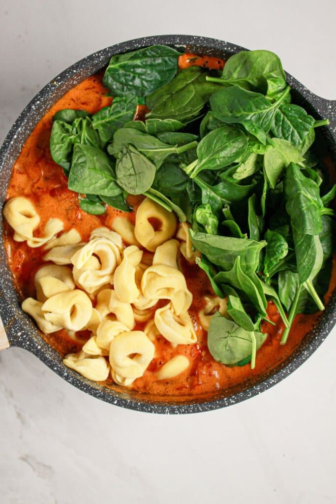 A black pan containing tomato sauce, fresh spinach leaves, and uncooked cheese tortellini on a white surface.