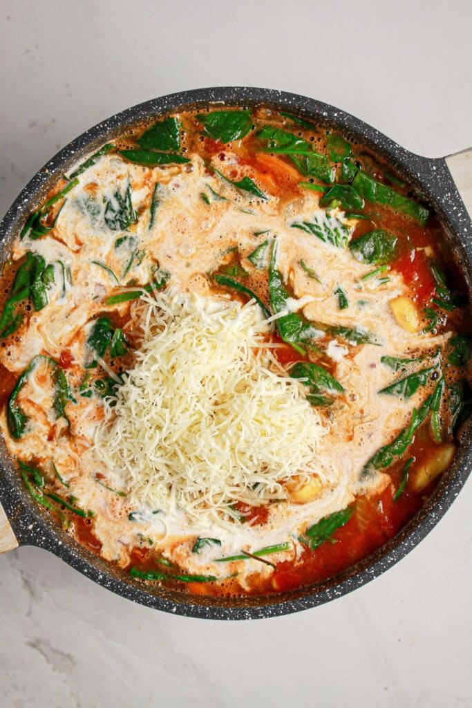 A pot of tomato-based soup with fresh spinach, grated cheese, and a swirl of cream on top, pictured from above.