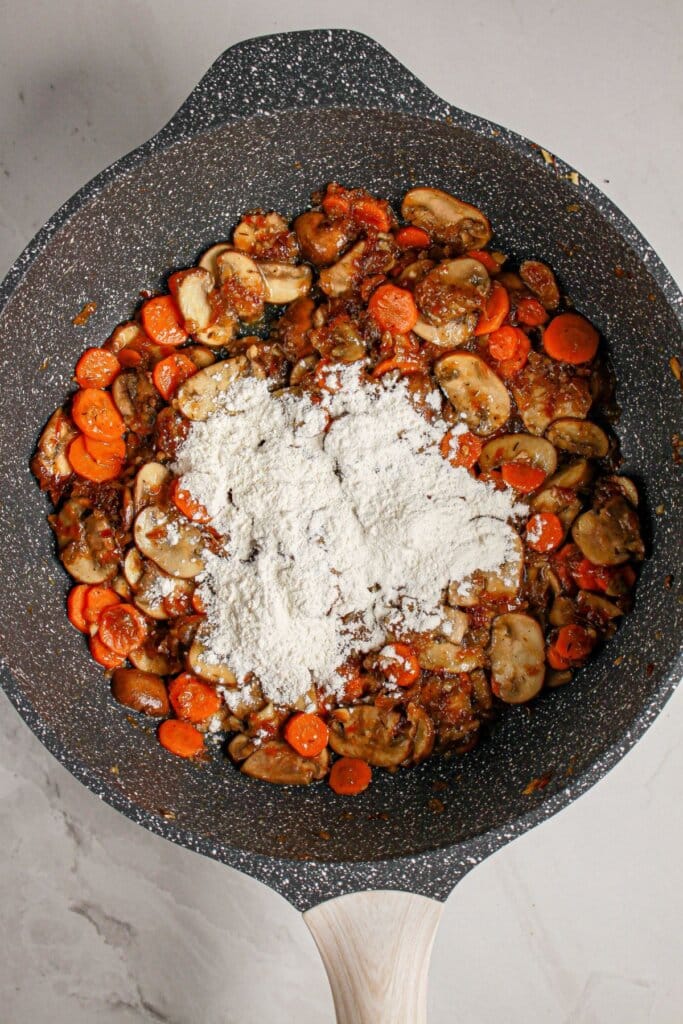 Flour being added to sauteed vegetables in a skillet.