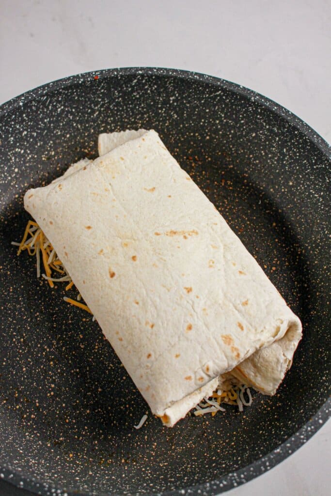 Rolled flour tortilla being cooked on a bed of shredded cheese in a skillet.