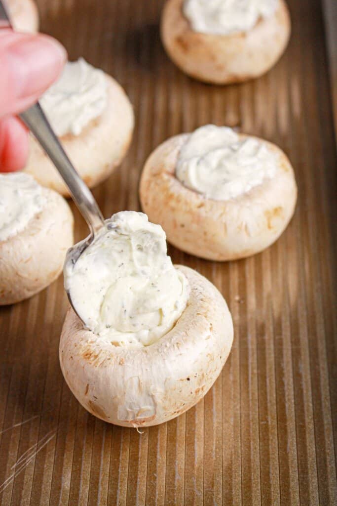 Cheese mixture being added to the center of mushroom on a baking dish.