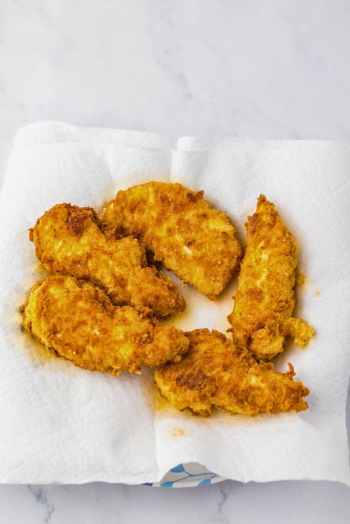 Fried chicken tenders drying on a paper towel lined plate.