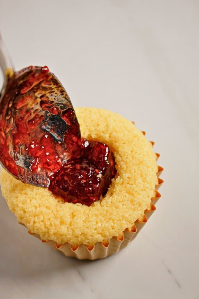 Raspberry jam being spooned into the center of a cupcake.