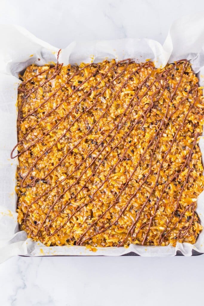 Chocolate being drizzled on top of dessert in a parchment lined baking pan.
