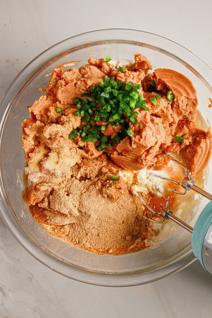 Refried beans, jalapeno, sour cream, taco seasoning, and cream cheese being mixed in a bowl.