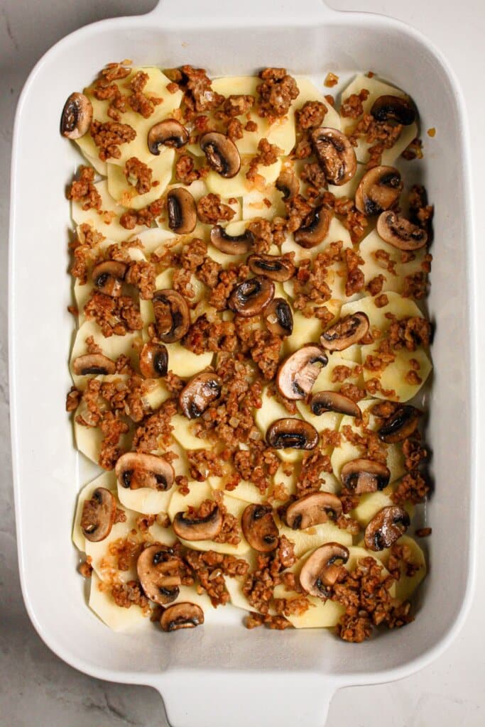 Mushrooms and cooked meat being sprinkled on top of a baking dish.