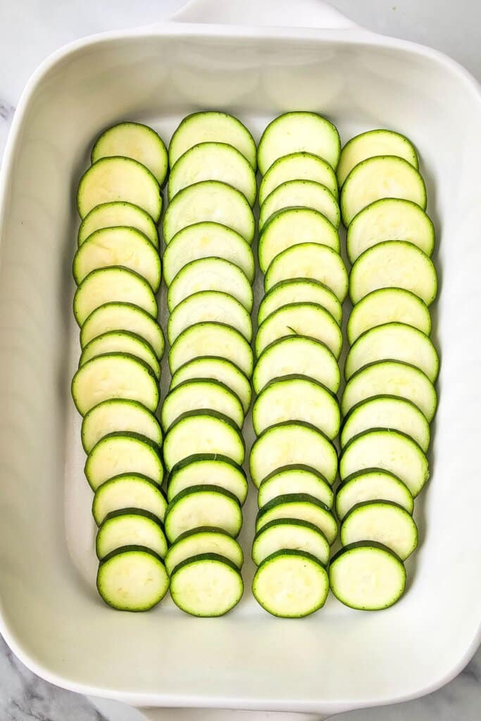 Slices of zucchini in the bottom of a white baking dish.