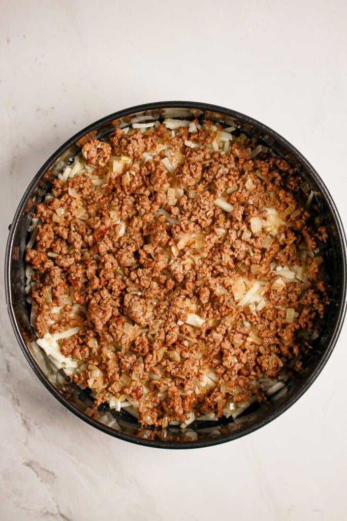 Beef being layered to top of hashbrowns in a springform pan.
