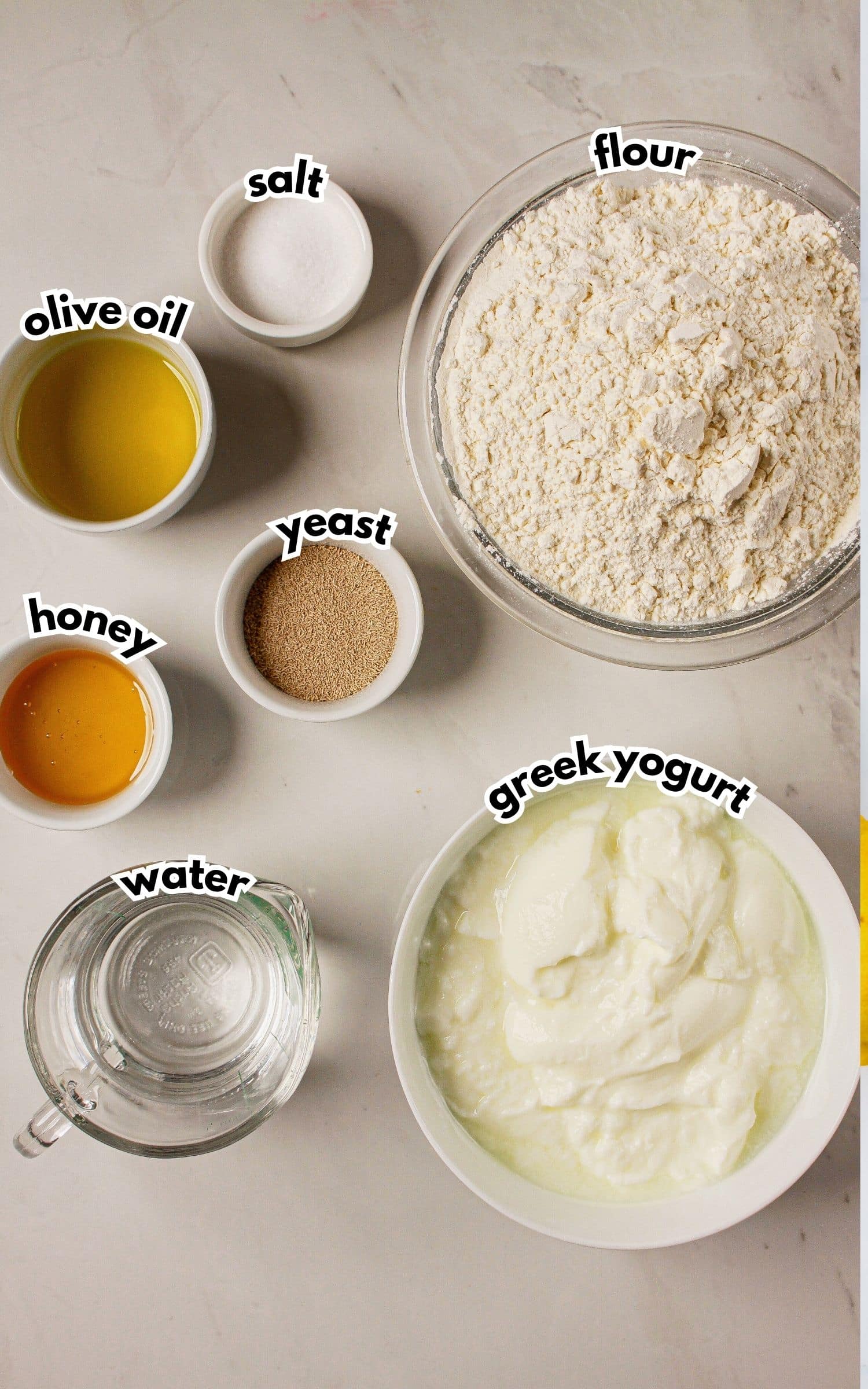 Bowls of salt, olive oil, honey, yeast, water, greek yogurt, and flour on a counter.