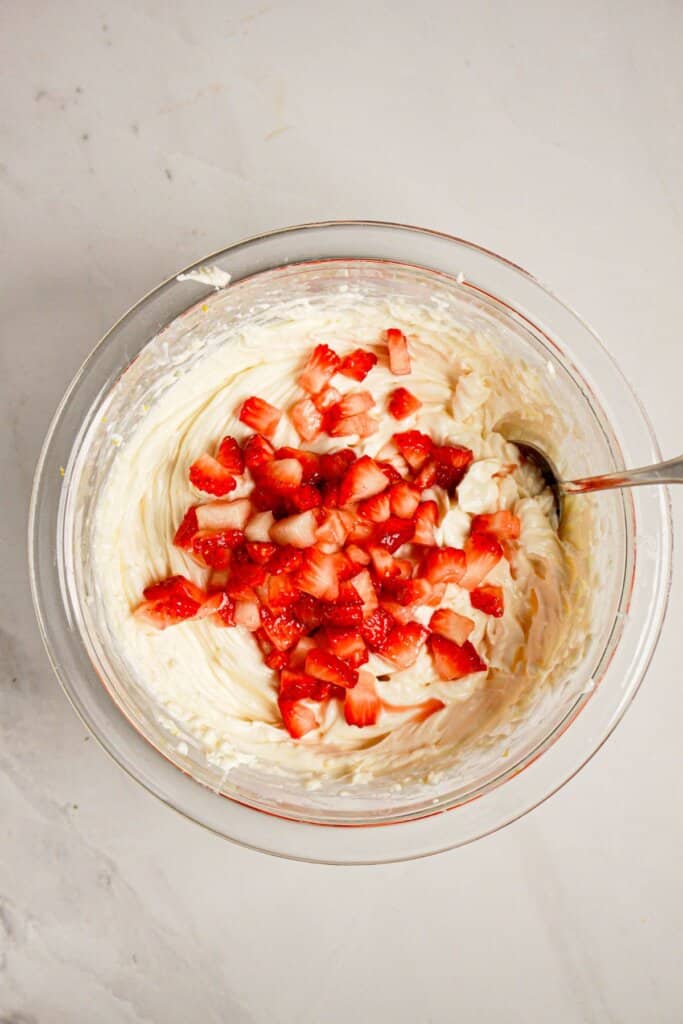 Fruit being stirred into cream cheese batter in a glass bowl.