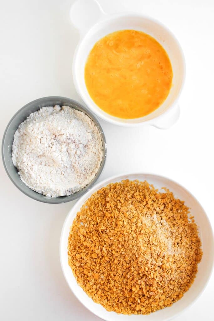 Bowls of egg, flour, and cornflakes on a counter.