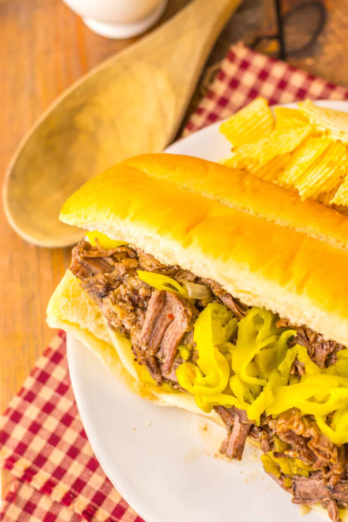 Overhead view of a slow cooker Italian beef sandwich with shredded beef and giardiniera peppers on a hoagie roll, served on a red checkered cloth.