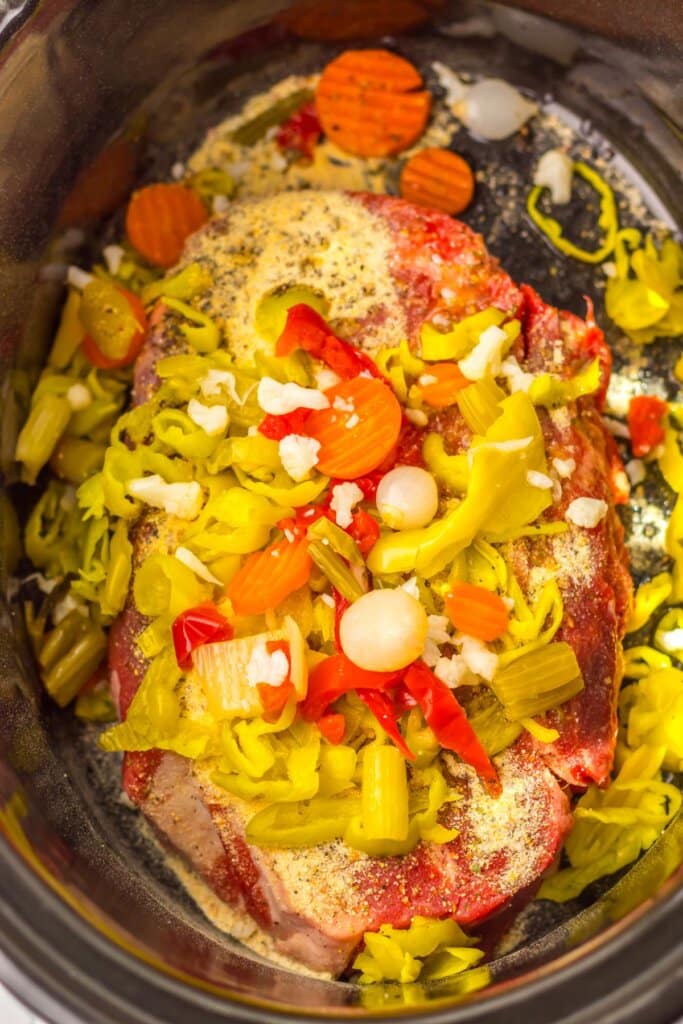 Raw chuck roast in a slow cooker surrounded by giardiniera, pepperoncinis, carrots, and seasonings before cooking.