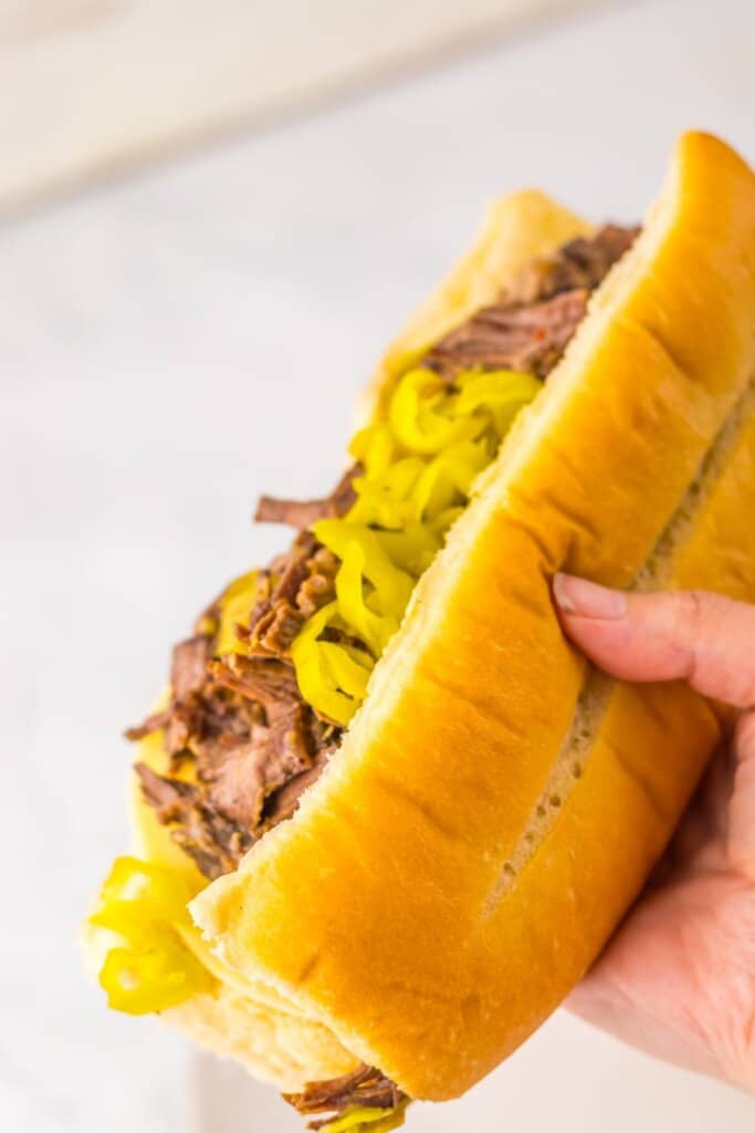 Hand holding a slow cooker Italian beef sandwich with giardiniera peppers on a hoagie roll.