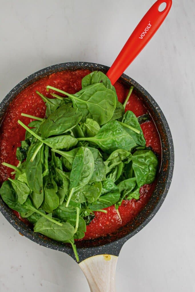 Spinach being added to sauce in a skillet.